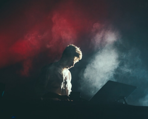 A musician in a white shirt on stage with white and red lights breaking through the smoke behind his back